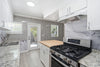 A modern kitchen interior with white cabinets, a stainless steel gas range, granite countertops, gray marble backsplash, and gray textured wall panels. A glass door is visible in the background.