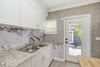 A modern kitchen interior with white cabinets, a double-bowl stainless steel sink, a marble-patterned backsplash, and a white door with a glass panel. The wall features vertical wood slats, and a wooden table edge is visible in the foreground.