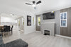 A modern living room with light wood flooring, a gray fireplace mantel, a mounted flat-screen TV, gray paneled walls, and an arched opening leading to a dining and kitchen area.