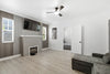 A modern living room with light wood flooring, white walls, a built-in gray fireplace with a mounted TV above it, a ceiling fan, and part of a gray sectional sofa in the foreground.